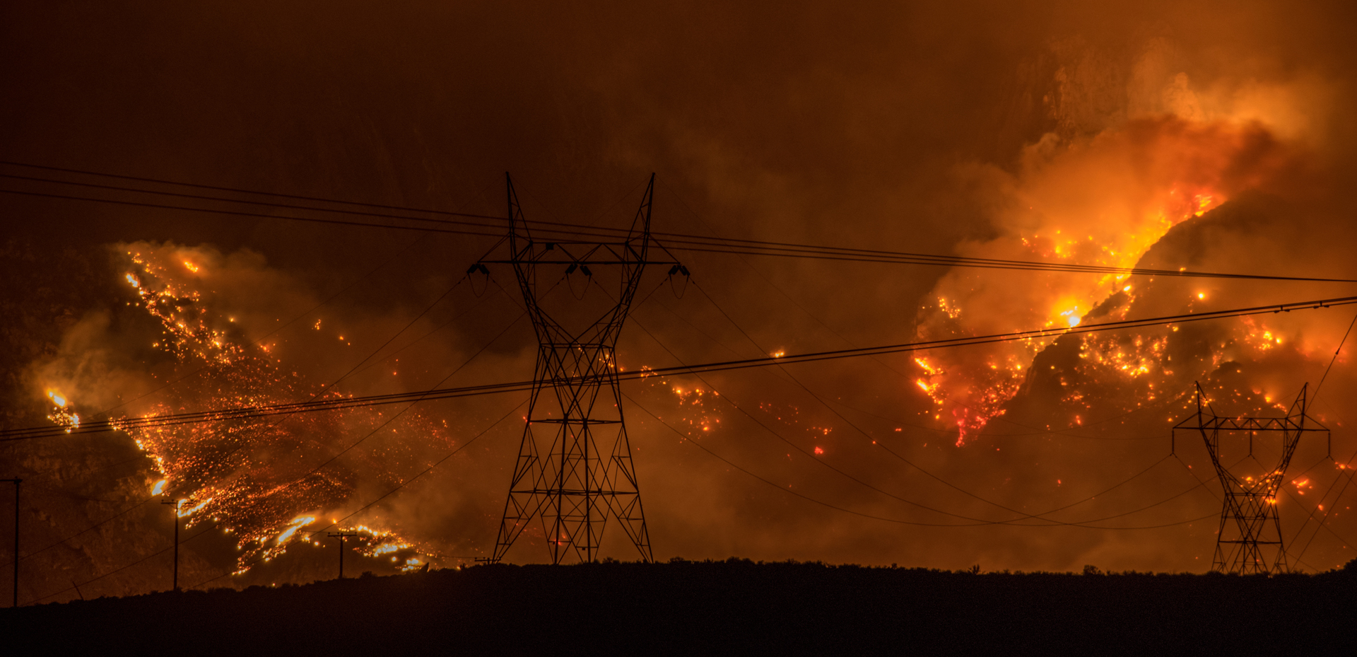 An intense wildfire, glowing orange is spreading in the background behind two tall electricity pylons