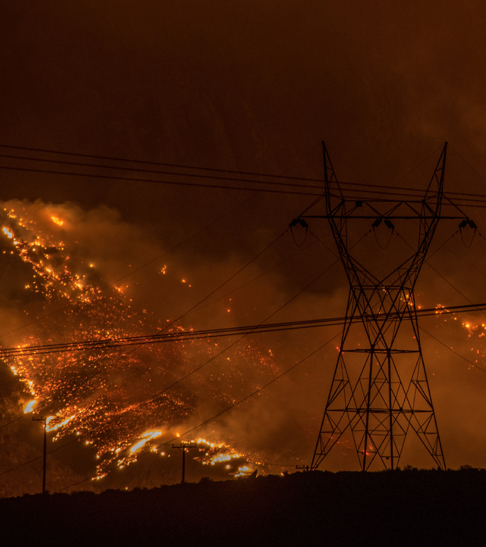 An intense wildfire is roaring behind two tall electricity pylons